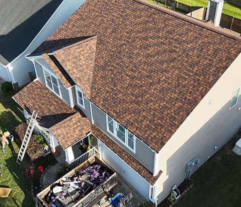 Aerial view of a two-story house with new brown asphalt shingles, a ladder leaning against the side, and equipment on the patio below.