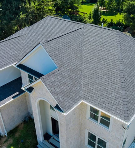 Aerial view of a house with light gray shingle roofing, white exterior walls, and multiple peaked roof sections, surrounded by green trees and grass.