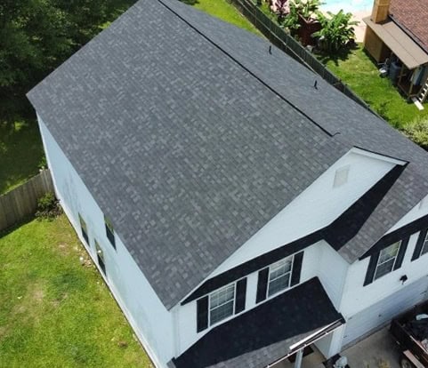 Aerial view of a two-story white house with a dark gray shingled roof and black window shutters, surrounded by a green lawn and a wooden fence.