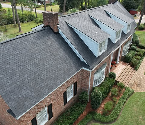 A large brick house with a dark gray shingle roof, dormer windows, white trim, and a landscaped front yard.
