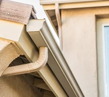 Close-up view of a beige rain gutter attached to the edge of a house roof, with a downspout leading water away from the structure.
