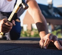Person kneeling on a roof, holding a nail in one hand and a hammer in the other, preparing to drive the nail into roofing material.