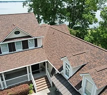 Aerial view of a house with brown shingle roofing, white trim, front porch, and steps, surrounded by trees and near a body of water.