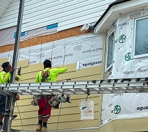 Two construction workers in safety gear install siding panels on the exterior wall of a house using tools, while standing on scaffolding.