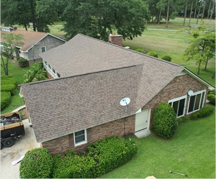 Aerial view of a single-story brick house with a brown shingle roof, surrounded by green lawns and trees.