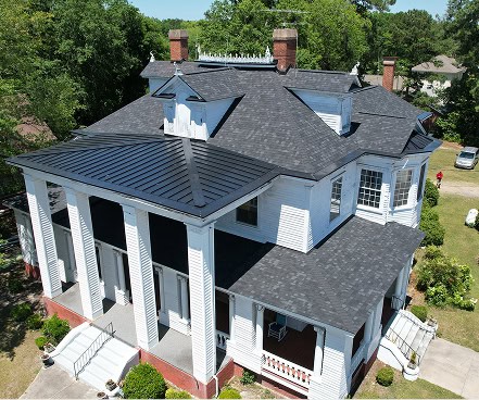 A large, two-story white house with black roofs, tall columns at the entrance, multiple chimneys, and surrounding greenery.