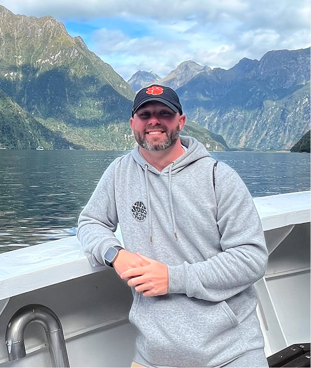 A man in a gray hoodie and cap stands on a boat with mountains and a lake in the background under a partly cloudy sky.