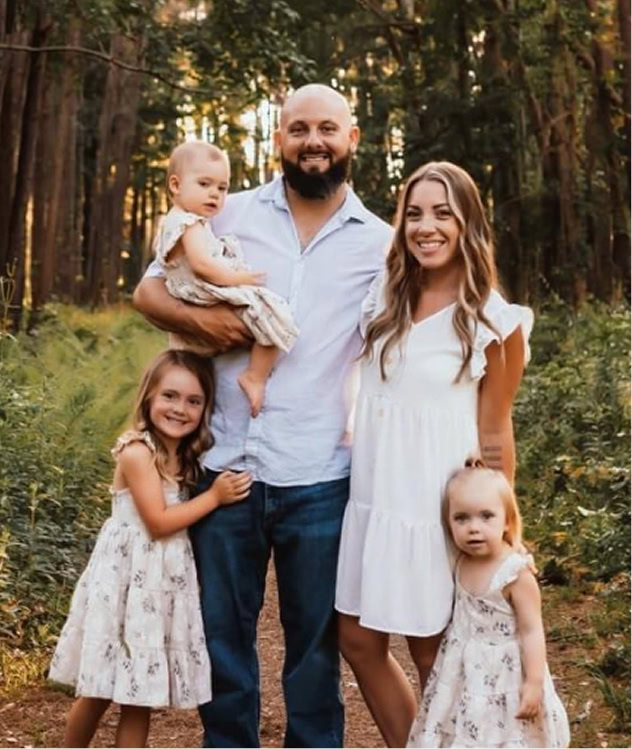 A man and woman stand on a wooded path with three young children. The man holds a baby; all are smiling and dressed in light-colored, summer clothing.
