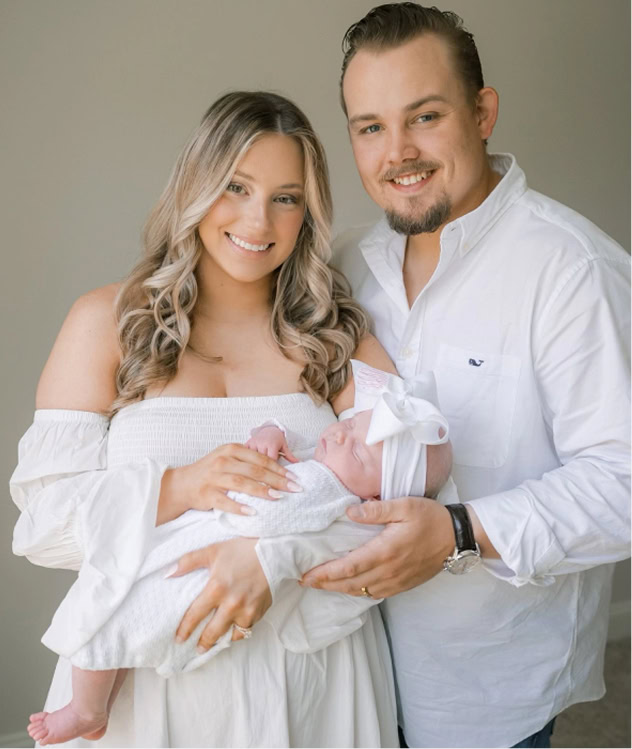 A woman and man, both wearing white, smile while holding a newborn baby dressed in white with a bow headband.