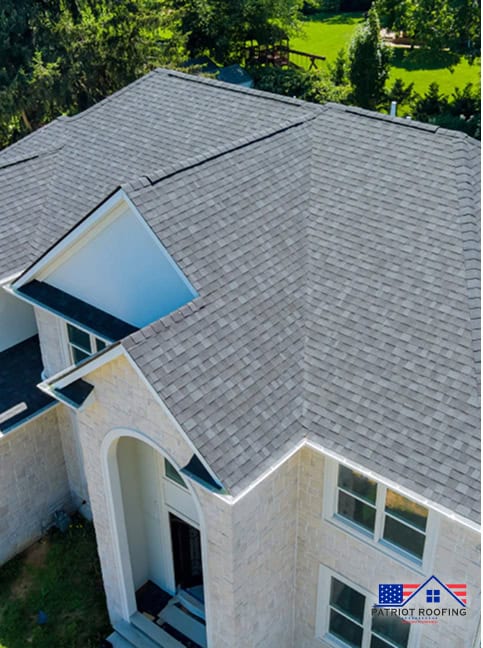 Aerial view of a house with light gray shingle roofing, featuring white trim and a curved entrance; Patriot Roofing logo in the corner.