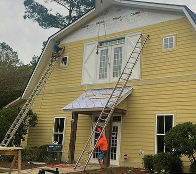Two workers use ladders to install or repair siding on the upper part of a yellow house, with construction materials and tools visible around the entrance.