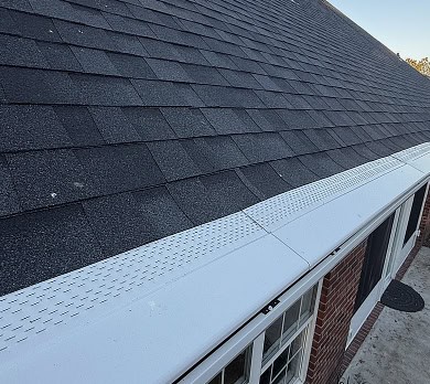 View of a house roof with dark asphalt shingles and a white gutter guard installed along the edge above windows and a brick wall.