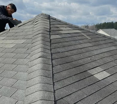 A person working on the roof of a house with gray asphalt shingles under a partly cloudy sky.