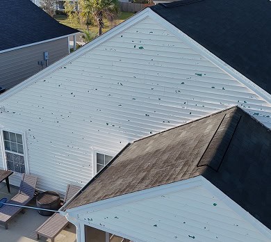 White house exterior with multiple green spots and dents on the siding, indicating damage. Patio furniture is visible on the ground near the wall.