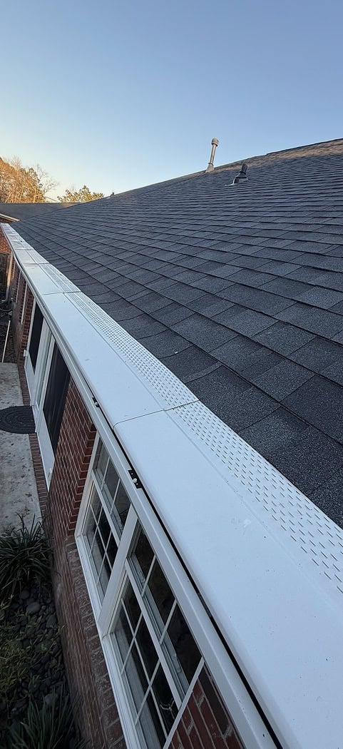 View of a sloped roof with dark shingles and a white gutter guard, above windows on a brick house under a clear sky.