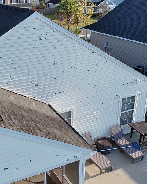 White house siding with numerous green paint chips and marks, next to a patio area with loungers, a table, and a grill; neighboring houses are visible in the background.
