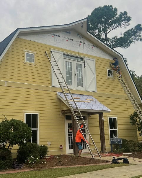 Two workers on ladders repair the siding and roof area of a yellow house, with tools and materials visible around the entrance.
