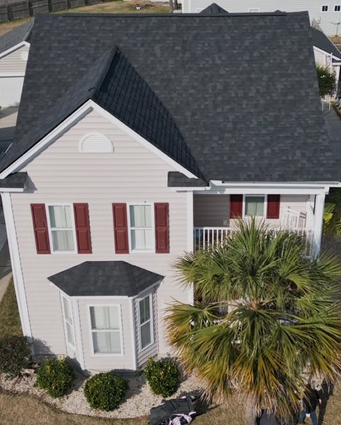 A two-story beige house with dark gray roof shingles, red shutters, white trim, and a small front yard featuring a palm tree and landscaping rocks.