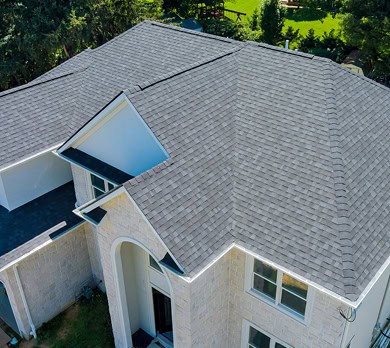 Aerial view of a house with a complex gray shingle roof, white siding, multiple roof peaks, and large windows.