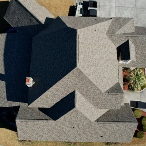 Aerial view of a house with a complex, multi-faceted gray shingle roof, visible chimney, driveway, and surrounding yard.