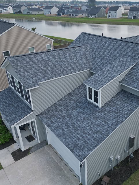 Aerial view of a gray house with asphalt shingle roofing in a suburban neighborhood near a pond.