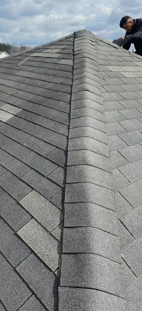 A person inspects or repairs the shingles on the ridge of a gray shingled roof under a partly cloudy sky.