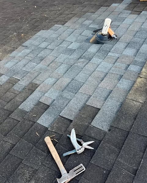 Partially replaced roof shingles with tools, including a hammer, pry bar, and pliers, lying on the shingles near a roof vent pipe.