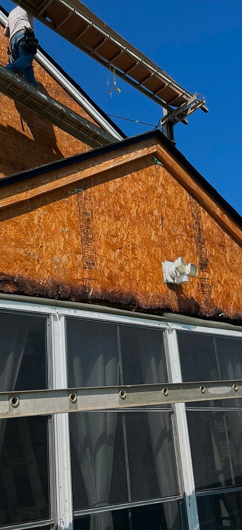 Person standing on a rooftop next to a ladder, with an unfinished wooden exterior wall and a motion-sensor floodlight attached. Blue sky in the background.