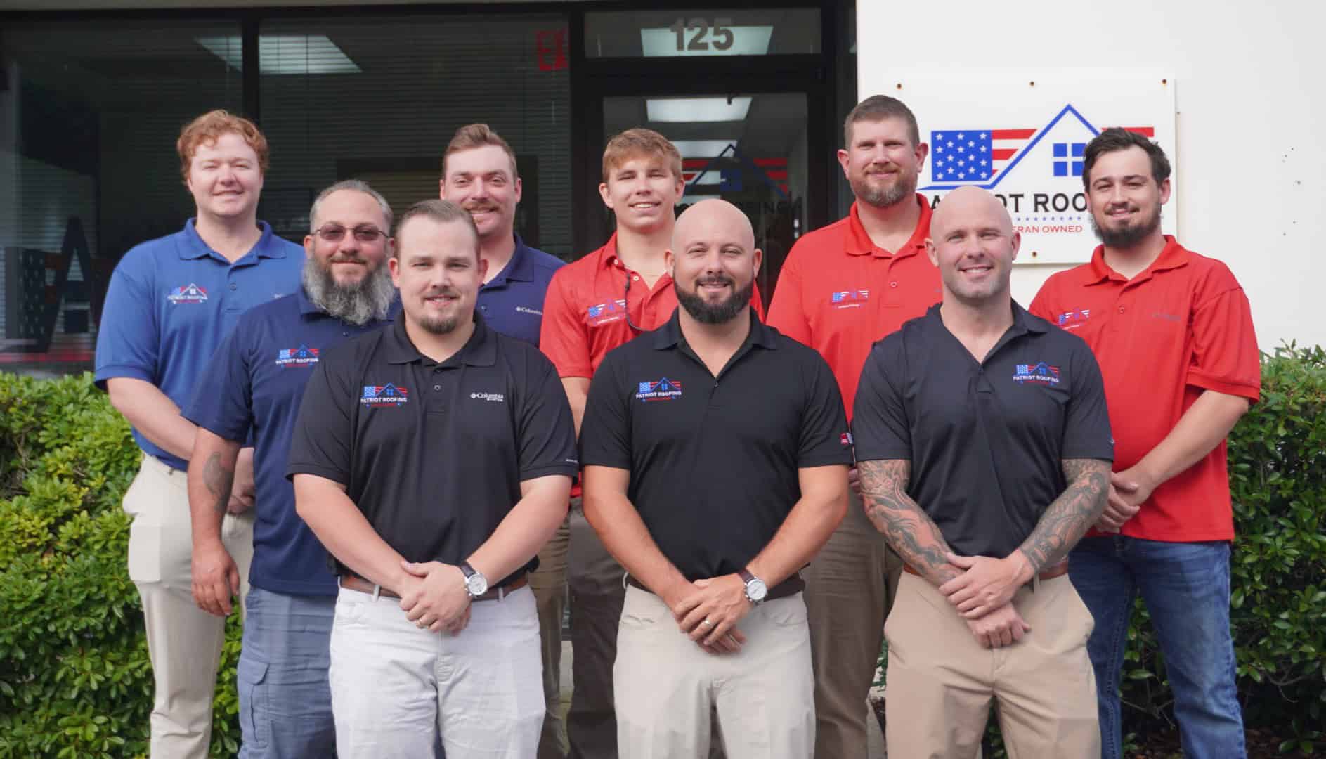 Nine men stand in two rows in front of a building entrance and a "Veteran Owned and Operated Roofing Company" sign, wearing matching red or black company shirts.