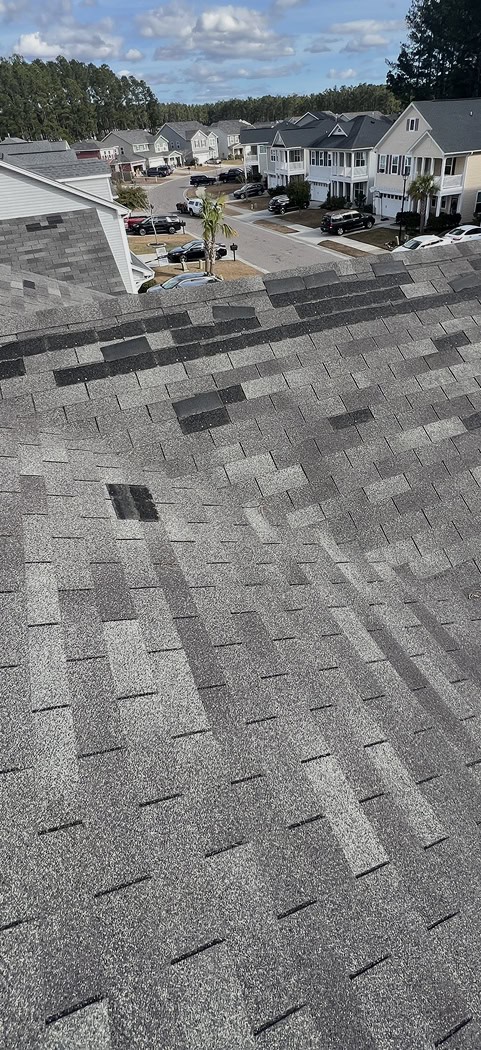 View of a residential neighborhood from a rooftop, showing asphalt shingles with one visibly different, darker patch among the uniform pattern.