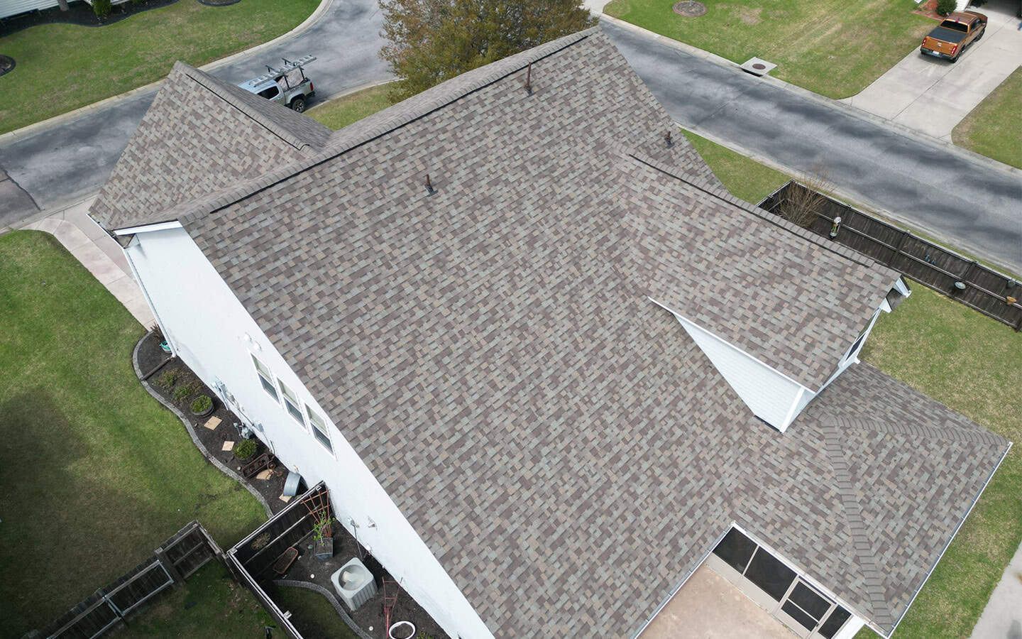 Aerial view of a residential house with a large, multi-gabled roof covered in brown asphalt shingles, surrounded by lawns and neighboring streets.