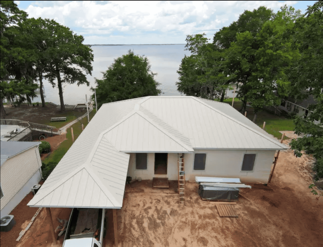 A single-story house with a metal roof is under construction near a lake, surrounded by trees and bare soil in the yard.