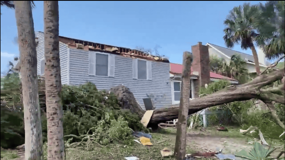 A large fallen tree lies on the ground in front of a house with roof damage; debris is scattered across the yard under a clear sky.